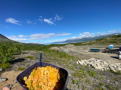 A close-up, over-the-shoulder view shows a dark purple rectangular container filled with pasta and vegetables. A fork is stuck into the pasta. The container rests on rocky, moss-covered ground. In the background, a bright blue sky with scattered white clouds stretches over a vast, mountainous landscape. A large body of water is visible in the distance, with snow-capped mountains beyond. A construction site is visible, with large piles of dirt and rocks, as well as several heavy-duty trucks, including a blue dump truck and a white semi-truck. A blue portable toilet sits near one of the trucks. The scene is likely outdoors in a natural, rugged environment, possibly a construction area near Kiruna, Sweden, given the mountainous terrain and the mention of the city.