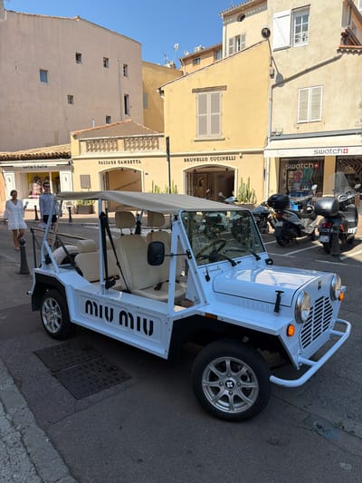 The image shows a white Miu Miu-branded vehicle parked on a street in Saint-Tropez, France. The vehicle, which appears to be a golf cart or a small electric car, is the main subject. It has a light-colored canopy, beige seats, and the "MIU MIU" logo printed on its side.

In the background, there are buildings with light-colored facades and shutters. Some of the buildings house shops, with visible signage including "Swatch," "Swatch+," "Brunello Cucinelli," "Passage Gambetta," and "Antik Batik." Two scooters are parked behind the Miu Miu vehicle.

Two people are visible in the foreground, standing on the street next to the vehicle. One person is wearing a white dress and the other a grey top and patterned skirt. They appear to be posing or interacting with the vehicle.

The scene is illuminated by what appears to be bright sunlight, suggesting it is daytime. The overall atmosphere seems relaxed and leisurely, typical of a vacation destination. The presence of luxury brands and the distinctive architecture of the buildings reinforce the Saint-Tropez location.