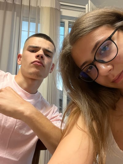 The image captures two individuals, one male and one female, in an indoor setting in Karavelovo, Bulgaria. The female is positioned to the right and closer to the camera, her face partially cropped on the right side. She has long, light brown hair that falls over her shoulders and obscures portions of the male. She wears black-framed eyeglasses with visible blue light reflections on the lenses, and her right eye is directed at the camera. Minor skin texture is visible on her forehead. The male is positioned to the left, slightly behind the female's shoulder and hair. He has a very short buzz cut and light stubble visible on his jawline. He wears a light pink crew-neck t-shirt with the words "Smiths Music" faintly visible in white text on the chest. His right arm is bent at the elbow, and his fist is lightly clenched and positioned in front of his body. Both individuals are looking directly at the camera. The background features interior windows draped with sheer white curtains, through which some daylight and hints of exterior blue are visible.