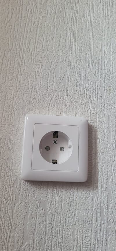 The image shows a white electrical outlet mounted on a textured white wall. The outlet is a European-style Schuko socket with two round holes and two grounding clips. The wall has a raised, linear texture, resembling wallpaper. The lighting is even, and there are no people or other objects visible in the frame. The context of Bulgakovo, Russia, is not visually evident from the image itself.