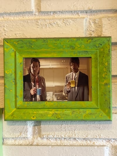 A framed photograph featuring two men in suits, each holding a mug, is displayed against a textured white brick wall. The photograph is contained within a bright green frame with yellow speckles. The man on the left has dark, shoulder-length hair and is looking directly at the viewer. He is wearing a dark suit, a white shirt, and a tie, with what appears to be a stain or blood splatter on his shirt and tie. He holds a blue mug in his right hand. The man on the right has curly dark hair and is looking slightly off to the side. He is also wearing a dark suit, a white shirt, and a tie, and holds a yellow mug in his right hand. The background of the photograph appears to be a kitchen or domestic setting, with cabinets and appliances visible. The overall presentation suggests a decorative item within an indoor space. The location is identified as Maracay, Venezuela.