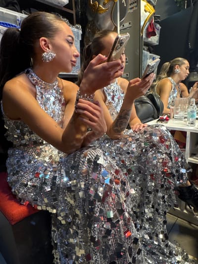 The image depicts an indoor scene, likely a backstage area or dressing room, located in دبي, United Arab Emirates. Two female individuals are seated, both wearing identical full-length, sleeveless halter-neck gowns constructed from numerous small, reflective silver squares.
The prominent individual in the foreground has dark hair styled in a high ponytail. She wears large, elaborate silver chandelier-style earrings and a wide, multi-tiered silver cuff bracelet on her left wrist. Her gaze is directed downwards at a smartphone held in both hands. A small tattoo is visible on her inner left forearm.
The second individual is positioned slightly behind and to the right, also seated, and is partially visible both directly and in a mirror reflection. She too holds a smartphone, directing her attention to its screen. On a white surface to her right, a black shoulder bag, a clear drinking glass, and a small bottle of water are present. The background is dimly lit and features various indistinct objects, including what appears to be a golden armored shoulder piece. Both individuals exhibit no direct interaction, focusing solely on their respective mobile devices.