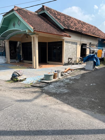 The image shows a residential house under construction in Kalitengah, Indonesia. The house has a red tile roof, a light-colored stucco exterior, and a light blue and green colored facade above the entrance. The entrance features two beige pillars supporting an arched roof decorated with a blue and white patterned trim. In front of the house, there is a paved walkway with colorful tiles. Scattered on the walkway are several pairs of sandals and a large, dark-colored, textured object resting on a rock. To the right of the walkway, a green plastic tub with a faucet is visible.

Adjacent to the house, on the right side of the frame, is a construction area. A stack of white bricks is covered with a blue tarp. There are also some wooden sawhorse-style supports and potted plants. Further to the right, laundry is hanging on a clothesline, with a bright yellow shirt prominently displayed. The ground in front of the house and the construction area is a mixture of dirt and gravel. A paved road runs in the foreground. The sky is blue with scattered white clouds.