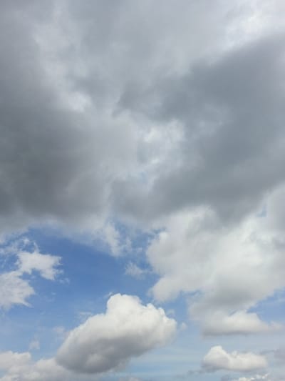 The image displays an overhead view of the sky above Ngọc Tụ, Vietnam. The upper and left portions of the frame are dominated by dense, dark grey overcast clouds. Towards the lower-middle and right-hand side, patches of clear blue sky are visible, interspersed with bright white, puffy cumulus clouds of varying sizes. No people, specific objects (other than clouds), actions, or interactions are present. The environment is atmospheric, with mixed cloud cover suggesting transitioning weather conditions.