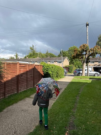 A child walks away from the camera, holding a clear umbrella with Marvel characters on it, down a gravel path. The child is wearing a dark green jacket and bright green trousers. The path is bordered by a wooden fence on the left and a grassy lawn on the right. In the background, there are houses, trees, and cars. A telephone pole with multiple wires extends upwards into a cloudy sky where a faint rainbow is visible. The location is Hazlemere, United Kingdom.