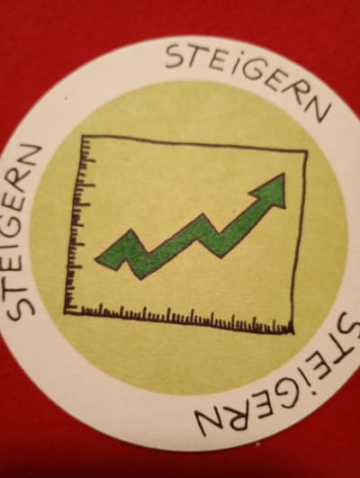 The image shows a circular sticker with a drawing of a stock chart on a yellow background. The sticker has the word "STEIGERN" written in black in an arc around the top and bottom edges, with "STEIGERN" also written vertically on the left side. The stock chart depicts an upward trending line graph with an arrow at the end, indicating growth. The sticker is placed on a red surface, possibly fabric. There are no people, actions, or interactions depicted. The location in the city of Rosenheim, Germany, is not discernible from the image itself.