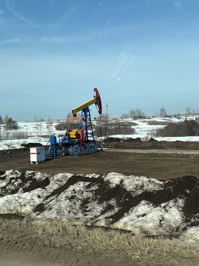The image shows an oil pump jack in a rural setting, likely located near Ustinovo, Russia. The pump jack is painted with red, yellow, blue, and black details, and is situated in a snowy field interspersed with bare trees and vegetation. The ground is a mix of brown earth and patches of melting snow. A utility box with red markings sits adjacent to the pump. The sky above is clear and blue. The scene appears to be taken during the daytime.
