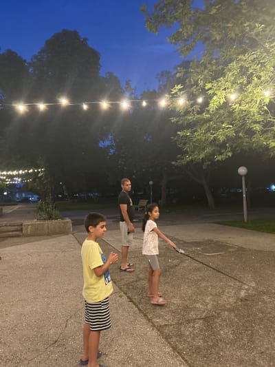 In the city of Parvomay, Bulgaria, a family is enjoying an evening outdoors. The scene captures a twilight ambiance, with a clear, dark blue sky overhead. String lights are visible, casting a warm glow across the area.
In the foreground, a young boy stands on a paved surface, wearing a yellow t-shirt and striped shorts. To his right, a young girl, dressed in a white patterned top and gray shorts, is holding a leash attached to an unseen dog, suggesting she is walking it. A man stands behind them, wearing a black t-shirt and light-colored shorts. He appears to be observing the children.
The background reveals a park-like setting with large trees. The ground is paved with concrete and stone, with some areas appearing damp. In the distance, streetlights and buildings are faintly visible, indicating an urban environment. The overall atmosphere is one of relaxed family time in an outdoor public space.