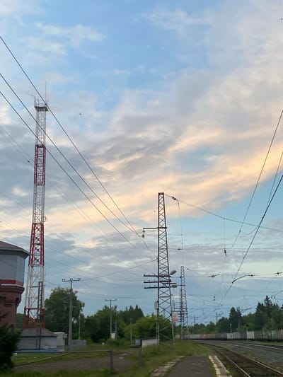 The image depicts a railway scene at what is identified as Borodulino, Russia. Dominating the foreground are railway tracks curving towards the right of the frame. Along the tracks, a platform is visible. To the left, a large water tower stands, adjacent to a tall, red and white lattice tower, likely a telecommunications mast. Several other similar lattice towers are visible in the distance, along with overhead power lines that crisscross the sky, indicating an electrified railway. A dense line of trees borders the railway on the right. The sky is filled with a mix of blue and white clouds, with hints of sunset colors. Several birds are visible in flight. The overall impression is of an industrial or semi-industrial area on the outskirts of a town.