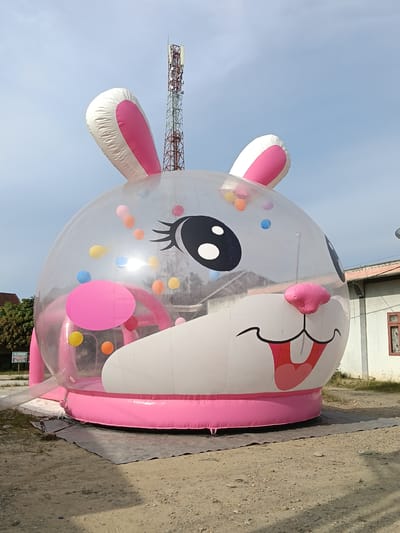 An inflatable, clear bunny head structure with pink ears is positioned on a dirt ground in Cempeudak, Indonesia. The bunny's face features large black eyes with white pupils, a pink nose, and a wide mouth with a pink tongue and red lips. Inside the clear portion of the bunny's head are several small, colorful balls in shades of pink, yellow, blue, and orange. A pink base supports the inflatable structure. Behind the bunny, a tall telecommunications tower stands against a blue sky with scattered clouds. To the right of the tower is a building with a white facade and a red-tiled roof. To the left of the bunny, a few trees are visible, and a sign with Indonesian text is mounted on a post. The ground is a mix of dirt and small stones, with some dry grass.