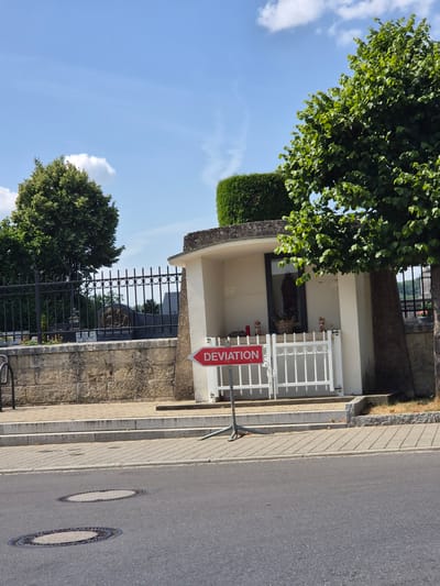 A tilted, eye-level, medium shot captures a street scene in Käerjeng, Luxembourg, on a bright, sunny day with scattered clouds. The foreground is dominated by an asphalt road with two manhole covers visible. A sidewalk made of light-colored paving stones runs alongside the road, bordered by a stone wall. Adjacent to the sidewalk, a building with a curved, light-colored facade features an open alcove containing a statue and a basket of flowers. A white metal gate is in front of this alcove. A red and white "DEVIATION" sign with an arrow pointing left is prominently displayed in front of the gate.
To the left of the building, a tall, dark metal fence encloses a grassy area with trees and more statues. Lush green trees with dense foliage partially obscure the sky and the building. The scene suggests a peaceful, everyday setting, possibly near a monument or a place of historical significance given the presence of statues and what appears to be a memorial or shrine within the building's alcove. The sunlight casts distinct shadows, indicating it is likely daytime, perhaps late morning or early afternoon. No people are visible in the frame. The visible text on the sign reads "DEVIATION".
