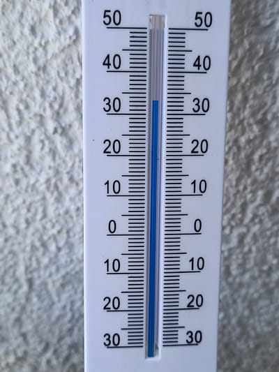 A close-up, eye-level shot shows a white thermometer mounted on a textured, light-colored wall. The thermometer has black markings and numbers indicating temperature in Celsius, ranging from 0 to 50 degrees. The blue liquid inside the thermometer indicates a temperature between 25 and 30 degrees Celsius. The thermometer is positioned vertically. The background is a stucco-like wall with visible texture and shadows. The image does not contain any people or specific city landmarks to determine its location in Oberviechtach, Germany.