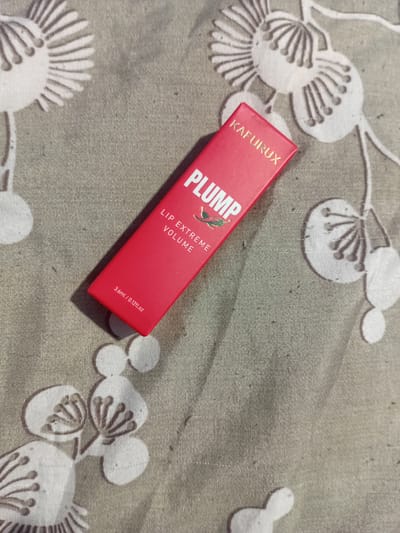 A red lipstick tube with the text "PLUMP LIP EXTREME VOLUME" and "KAFURUX" is lying on a grey fabric with a white floral pattern. The tube is slightly angled, with the top left corner pointing towards the top left of the image. The fabric appears to be a tablecloth or a similar textile. The lighting suggests it is indoors. There is no indication of the location being in Bocani, Moldova.
