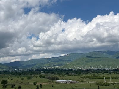 The image depicts a panoramic view of the city of Sagarejo, Georgia, nestled in a valley at the base of lush green mountains. The sky above is a vibrant blue, punctuated by dramatic white cumulus clouds, suggesting fair weather with a possibility of scattered showers.
In the mid-ground, Sagarejo appears as a sprawling settlement with numerous buildings, characterized by a mix of residential houses and some larger structures. The city is spread out, with trees and greenery interspersed throughout. Dominating the hillside behind the city, a large inscription is visible, appearing to be the name of the city in a national flag's colors.
The foreground consists of rolling green fields, dotted with scattered trees and some agricultural land. A simple, light-colored building or structure, possibly a farm building, is situated in the middle of the field. Fencing is visible in sections, and power poles line a dirt road or path that cuts through the landscape. The overall impression is one of a rural or semi-rural setting in a picturesque mountainous region. There are no people visible in the image.