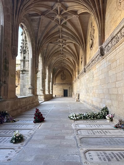 The image depicts an interior cloister of the Santiago de Compostela Cathedral in Santiago De Compostela, Spain. The cloister features Gothic architecture with ribbed vaults and stone columns. Along the right wall, there are several stone tombs with inscriptions and floral arrangements, indicating a burial site within the cloister. The floor is paved with stone. The lighting suggests it is daytime, with sunlight casting shadows and highlighting the architectural details and the tombs. No people are visible in the scene.