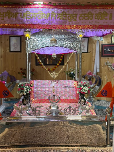 This image shows the interior of a Sikh Gurdwara, likely located in Chumkar, India. The scene is dominated by a raised platform covered in ornate pink and gold fabric, on which rests a large, silver-colored structure resembling a canopy. Beneath the canopy, a book, likely the Guru Granth Sahib, is covered with a patterned cloth.
Various religious symbols are visible, including the Khanda, a Sikh emblem, depicted in silver on the platform and as part of a flag. Two orange flags with Khanda symbols are placed on either side of the platform. Decorative elements include floral arrangements in vases, silver eagles, and metallic finials.
The background consists of wooden paneled walls, adorned with framed pictures and religious texts. A purple and gold banner with Sikh inscriptions hangs across the top of the scene. The lighting appears to be from ceiling-mounted fixtures. There are no people visible in the image, and the scene is static, suggesting a devotional space prepared for religious observance.