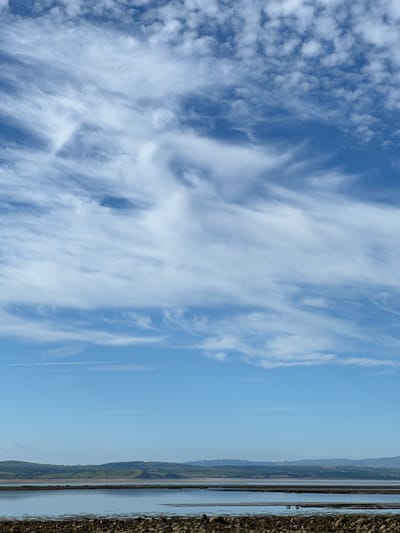 This media file captures a scenic view of a coastal landscape under a bright, blue sky with scattered white clouds. The image is oriented vertically, with the sky occupying the left two-thirds and the land and sea on the right.
The sky is filled with wispy, streaky clouds, particularly on the left side, creating a dynamic and textured appearance. Towards the right, the sky transitions to a clear, lighter blue.
The right side of the image features a calm body of water reflecting the sky, with a distant shoreline visible. The shoreline is characterized by rolling hills and fields, interspersed with what appear to be wind turbines, suggesting a wind farm in the area. The far right edge of the image shows a rough, rocky embankment or shoreline, adding a contrasting texture to the smooth water and sky. There are no discernible people or specific objects that suggest a particular activity. The lighting indicates it is daytime, likely a clear and sunny day. The presence of wind turbines and the coastal setting suggest this is a rural or semi-rural location, possibly along the coast of Ireland, given the location context.