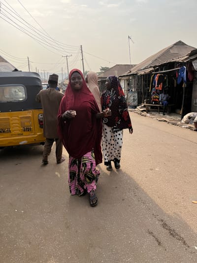Three women walk on a street. One woman wears a burgundy headscarf and pink patterned skirt. The second wears a beige headscarf and is partly visible. The third wears a dark floral headscarf and a white skirt with black polka dots. A man in a brown outfit and dark hat is partially visible to the left, standing by a yellow auto rickshaw. In the background, a building with clothes displayed outside is visible. Overhead wires stretch above the street.