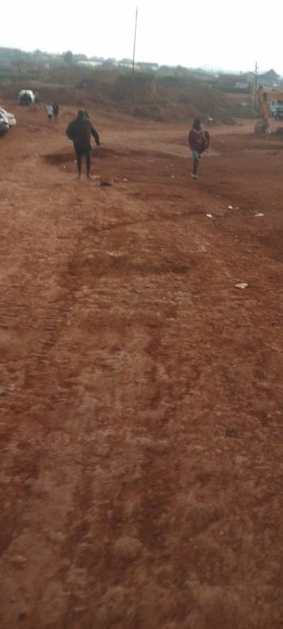 This media file captures a scene in Reifild-Ist, Nigeria, depicting a dirt and gravel terrain under an overcast sky, suggesting either dawn, dusk, or a cloudy day. In the mid-ground, two individuals are walking away from the viewer. The person on the left appears to be wearing a dark hooded jacket and dark pants. To their right, another person is visible, dressed in a red jacket or backpack and light-colored pants.
Further in the background, a few other people are present, appearing smaller due to distance, and some vehicles are parked on the left side of the frame. On the right side of the background, construction equipment, including what looks like a yellow excavator, is visible, hinting at ongoing development or infrastructure work in the area. The landscape is gently sloped and appears somewhat arid, with sparse vegetation visible on the distant hills. The overall impression is a utilitarian or developing environment. There is no visible text in the image.