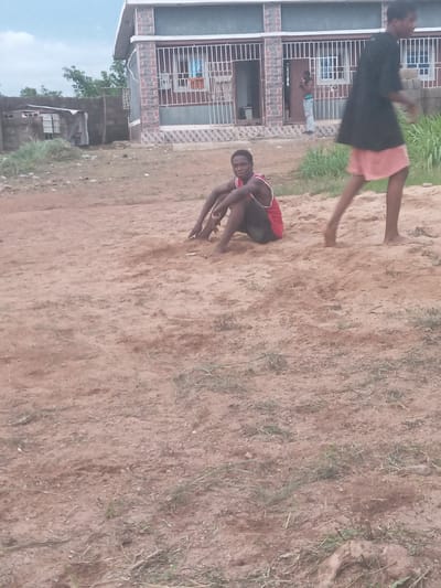 The image shows a dusty outdoor area in Achara Ndiagu, Nigeria. A man wearing a red-trimmed tank top and dark shorts is crouched down, appearing to be playing with the dirt or examining something on the ground. A second person, partially visible, is wearing a dark top and light-colored shorts and appears to be walking or running past in front of a building. The building is a multi-story structure with barred windows and a brick exterior. Another person can be seen in the doorway of the building. The ground is bare earth with sparse patches of grass. In the background, there are trees and other buildings visible.