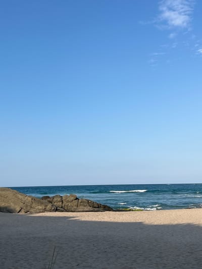 The image depicts a serene beach scene in Ahtopol, Bulgaria. A clear, bright blue sky dominates the upper portion of the frame. Beneath it, the deep blue sea meets the shore with gentle waves. A line of large, dark rocks juts out into the water from the left side of the image, providing a rugged contrast to the soft, light-colored sand of the beach in the foreground. The sand is smooth and shows subtle undulations from wind or water. The overall impression is one of tranquility and natural beauty, characteristic of Ahtopol's coastline. There are no people visible in the image, emphasizing the peacefulness of the location.