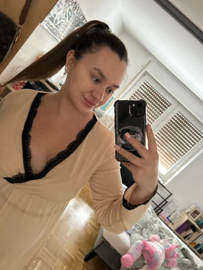 A woman with her hair in a high ponytail takes a selfie in a room. She is wearing a pale peach-colored dress with black lace trim along the neckline and sleeves. The dress has a deep V-neck. She is holding a black smartphone in her right hand. In the background, there is a window with horizontal blinds, a shelf unit filled with books and decorative items, and a bed with a colorful plush toy. The floor is made of light-colored wood. The overall impression is of a domestic setting, likely a bedroom or living area. This image is not sufficient to determine the location in the city of Waldbröl, Germany.