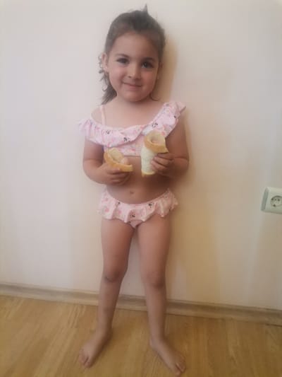A young girl is standing in front of a light-colored wall, wearing a pink bikini with a floral and Mickey Mouse print. She is holding two pieces of bread, one in each hand, and appears to be smiling at the camera. The girl has dark hair pulled back and is barefoot. The floor is made of light-colored wood. In the background, on the right side of the image, there is a white electrical outlet on the wall. The scene appears to be indoors, possibly in a home. The lighting suggests it could be daytime, but no specific time can be determined. The overall mood of the image is cheerful and candid. There is no visible text in the image.