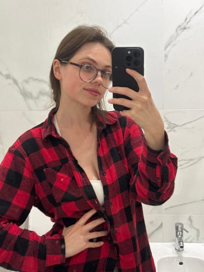 A young adult female is depicted taking a mirror selfie in an indoor setting, likely a bathroom, characterized by white marble-patterned tiled walls and a white ceramic sink with a chrome faucet. The specific location within Scafati, Italy, cannot be determined from the visual information provided. The female has brown hair, styled down, and wears round, dark-framed eyeglasses. She is dressed in an unbuttoned red and black buffalo plaid long-sleeved shirt over a white tank top or bralette. Her right hand holds a black smartphone with a dark case and a visible multi-lens camera array, positioned to capture her reflection. Her left hand rests on her midriff, with fingers splayed. Her gaze is directed towards the smartphone camera in the reflection, and her expression is neutral with a slight smile.