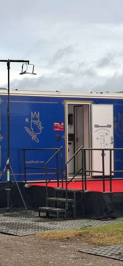 A blue portable restroom trailer with ornate gold detailing is parked outdoors. The trailer has a red carpet leading up to its entrance, which is marked by a white door with the text "luxury toilets" and a drawing of a toilet. A metal staircase with railings provides access to the door. The side of the trailer features a graphic of a crown and a sword, along with the text "royalflush.ee". Black fabric covers are visible on the ground next to the trailer, and a metal structure resembling a scaffolding or portable stage is set up in front of it. The ground is paved with interlocking stones. In the background, a cloudy sky suggests an overcast day. The image appears to be taken in Kuressaare, Estonia, possibly during an outdoor event.
