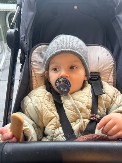 An infant is depicted seated in a dark-colored stroller, positioned outdoors in Longone al Segrino, Italy. The child, appearing to be a baby or young toddler, wears a gray knitted beanie and a quilted, light beige jacket. A blue pacifier with a white floral pattern is present in the infant's mouth. The infant's light-colored eyes are directed towards the left side of the frame. The left hand holds a light brown, elongated biscuit. The stroller features a padded insert with a faint geometric pattern and black harness straps, one of which has a patterned fabric piece. The background is out of focus, showing indistinct metal elements consistent with outdoor furniture, such as table and chair legs.