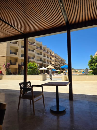 The image depicts a scene in Al Ahia', Egypt, looking out from under a slatted wooden structure. The view is of a multi-story hotel complex with sandy-colored walls and multiple balconies. A swimming pool area is visible with blue umbrellas and lounge chairs. A person is walking in the pool area. In the foreground, inside the slatted structure, there is a table and a chair. The floor is a light-colored stone or tile. The sky is clear and blue.