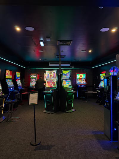 The image captures an indoor scene, likely a gaming arcade or casino, filled with numerous electronic gaming machines. The setting is dimly lit with artificial lighting, creating a vibrant and atmospheric environment primarily through the glowing screens and LED accents of the machines.
On the left side of the frame, a dark wall extends from the foreground into the background. Along its upper edge, a bright aqua-colored LED strip runs horizontally, casting a soft glow. Various light fixtures are mounted on this wall: several circular recessed lights, some emitting a warm yellow light, others a cooler white; and a couple of rectangular surface-mounted lights, one white and one with a red hue. A white rectangular air conditioning unit is also visible on the wall, along with a square recessed decorative element featuring a ribbed pattern.
The central and right portions of the image are dominated by rows of electronic gaming machines. These machines have multiple brightly lit screens displaying colorful graphics from various games. The casings of the machines are dark, often accented with vibrant LED strips in green, blue, and red, adding to the dynamic lighting of the space. In the mid-ground, a person is seated with their back to the viewer, actively engaged with a dual-screen gaming machine. This machine, like others, features glowing green LED strips along its base. Behind and around this individual, more machines stretch into the background, some with blue LED lighting.
The floor is carpeted with a subtle, tightly woven dark grey pattern. In the foreground, to the right of the person, a tall, black stand holds a white sign. The text on this sign is clearly legible and reads: "STAY SAFE. Gambling Helps When You Need It. If you or someone you know has a gambling problem, Help is available. Call 1-800-522-4700. National Council on Problem Gambling. www.ncpgambling.org". In the upper right corner, a dark blue LED strip marks another wall boundary, and a gray door or panel is partially visible. The overall impression is one of an active, modern entertainment venue focused on electronic gaming.