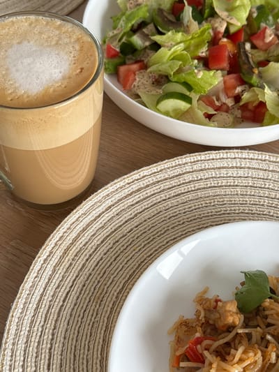 A close-up, high-angle shot shows a meal set on a wooden table. In the foreground, a white plate with rice and chicken is partially visible, placed on a beige woven placemat. To the left, a glass of coffee with foam is positioned next to the placemat. In the background, a white bowl filled with a salad of lettuce, tomatoes, and cucumbers, dressed with a creamy sauce, is visible. The setting appears to be a domestic environment, possibly a home in Гамово, Russia.