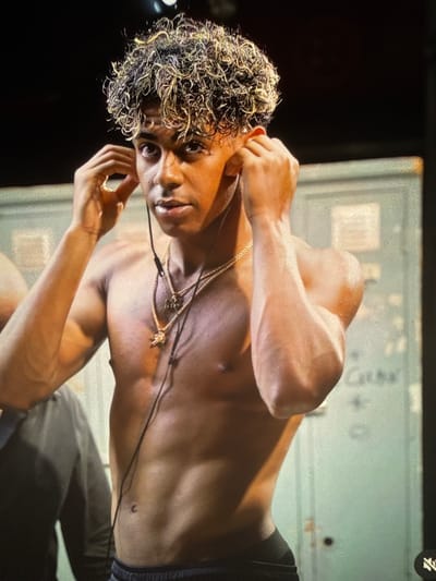 A young shirtless man with curly, blonde-streaked hair is shown from the chest up, putting earbuds into his ears. He is wearing two gold necklaces, one with a pendant and another with letters. The background appears to be a locker room with light green metal lockers. The lighting is dramatic, with some areas brightly lit and others in shadow. There is no indication of the location being Ravno pole, Bulgaria.