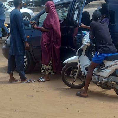 In Sardauna, Nigeria, a man in blue shorts and a black shirt sits astride a white motorcycle next to a dark blue car, leaning inside through the driver's side window. A woman in a maroon robe stands by the open passenger side door of the same car. To her left, a man in a dark blue suit holds a black object connected by a wire to the car. Other cars are visible behind the blue car. The ground appears to be a sandy or dirt surface.