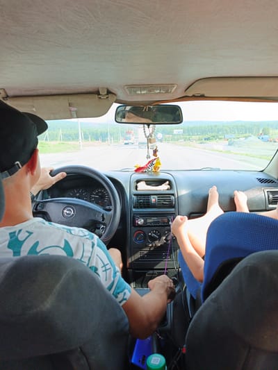 The image shows the interior of a car from the perspective of the front passenger seat, looking forward. A man wearing a black baseball cap and a white and teal patterned t-shirt is driving. His hands are on the steering wheel. In the front passenger area, a person's legs are propped up on the dashboard, wearing blue shorts. The car is on a road with trees and buildings visible in the distance. The sun is shining, indicating a daytime scene. The license plate of the car is not visible. The image does not provide specific information that would allow for a precise location within Nizhnii Atlyan, Russia, other than it is a road in or near a populated area.