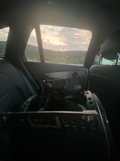 Inside a car, a dark-colored pet carrier sits on the back seat. The carrier has numerous ventilation holes and a metal frame. A dark-colored animal, likely a dog, is visible inside the carrier, with some of its fur peeking through the holes. The car's interior is dimly lit, with the focus on the carrier and its occupant. Through the rear windows, a sunset is visible, casting a warm glow on the cloudy sky and the distant, hilly landscape. The scene appears to be a still moment during a car journey, possibly in or near Donje Mrke, Montenegro, given the arid, mountainous terrain suggested by the view outside.