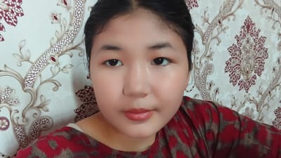 This is a close-up, waist-up photograph of a young woman with dark hair pulled back. She is looking directly at the camera with a neutral expression. Her skin is fair with a slight rosy blush on her cheeks. Her eyes are dark and almond-shaped, and she has subtle eye makeup. Her lips are painted a soft, peachy-pink color. She is wearing a red and gray patterned shirt. The background is a patterned wallpaper with floral motifs in shades of white, gold, and dark red. The lighting suggests it is daytime, likely indoors. The overall impression is a casual, personal photograph, possibly a selfie. The location context is Uchkun, Kyrgyzstan.