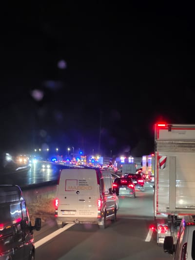 The image captures a nighttime traffic jam on a highway, likely near Uettingen, Germany, indicated by the presence of German license plates and street names on one of the vehicles. The road surface appears wet, suggesting recent rain or damp conditions, and is illuminated by headlights and streetlights, creating reflections. Several vehicles are stopped or moving slowly, including vans, cars, and large trucks. Emergency vehicle lights, possibly blue and red, are visible in the distance, hinting at an incident. One white van in the foreground has markings that read "MALEREI ANSTRICH SANIERUNG MABEKO" and "www.mabeko.at," identifying it as a painting and renovation service company. Another white truck in the middle ground has "KRONE" and "Cool Liner" branding. The scene conveys a sense of congestion and potential disruption on the highway at night.