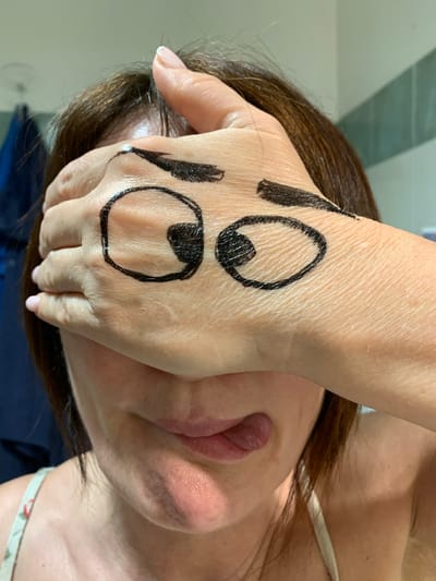 This media file features a close-up shot of a person's hand with cartoonish eyes drawn on it, obscuring another person's face. The hand is positioned over the face of the second person, with the drawn eyes facing the camera. The person whose face is obscured has brown hair that partially covers their face. The drawing on the hand consists of two large, circular eyes with black pupils. Above the eyes, there are two vertical black lines, resembling eyelashes or eyebrows. The skin on the hand shows visible pores and texture. The visible part of the obscured person's face includes their lips, which are slightly parted, and some of their chin and cheek. A strap from a piece of clothing, possibly a top, is visible near the neck area of the obscured person. The background is softly blurred, suggesting an indoor setting with neutral lighting. The overall impression is playful or humorous, due to the drawn-on eyes. There is no visible text in the image. The setting appears to be a casual, personal environment.
