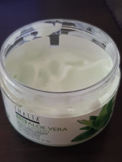 The image shows an open jar of Thalia Aloe Vera Skin Care Cream. The cream is a pale green color and has a smooth, whipped texture. The jar is made of clear plastic and has a white label with green text. The label indicates that the product is "REPAIR & HYDRATE ALOE VERA SKIN CARE CREAM" and contains "95% ALOE VERA." There are also images of aloe vera leaves on the label. The jar is sitting on a dark surface, possibly a table, and the background is out of focus and appears to be a dark color. There are no people or animals visible in the image. The location in the city of Sambir, Ukraine, is not depicted or implied in the image.