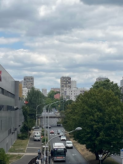 This image captures a street scene in Créteil, France, under a cloudy sky. In the foreground, a modern grey building stands to the left, with its facade featuring horizontal windows and some signage. A street runs diagonally through the middle of the frame, bordered by trees and sidewalks. Several vehicles are visible on the street, including cars, vans, and a bus with the number 217 displayed. There are streetlights and pedestrian crossings along the road. In the middle ground, a large, dense green tree dominates the right side of the frame. Beyond the trees, apartment buildings of varying heights, typical of urban residential areas, are visible against the overcast sky. The overall impression is of a residential urban environment.