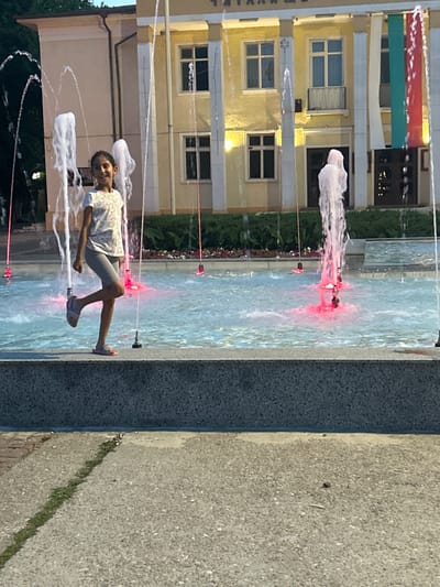 A young girl with dark hair, wearing a white t-shirt with a floral pattern and grey shorts, is standing in a fountain. She is looking towards the camera and smiling, with her left leg bent at the knee and her right foot resting on the fountain's edge. The fountain has multiple water jets, some illuminated with red lights, creating a dynamic visual. Behind the fountain, a building with a light-colored facade and several windows is visible. Vertical banners in white, green, and red hang from the building, suggesting a national or regional affiliation. The environment appears to be an outdoor public space, likely a town square or plaza, in Parvomay, Bulgaria. The ground in front of the fountain is paved with textured material, with a strip of grass running through it.