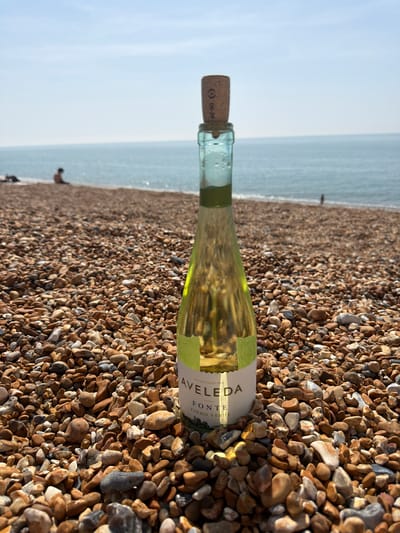 The image shows a bottle of Avelada Vinho Verde wine lying on a pebble beach. The bottle is oriented horizontally with the cork facing left and the label facing up. In the background, the sea and sky are visible with a few indistinct figures on the beach near the water. The pebbles are predominantly brown and grey, varying in size and shape. The location is likely a beach within the Folkestone and Hythe District, United Kingdom.