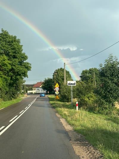A bright rainbow arcs across a cloudy, pale blue sky, with the end of the rainbow seemingly disappearing behind trees on the right side of the frame. In the foreground, a two-lane asphalt road with white double solid lines marking the lanes stretches into the distance. The road is wet, suggesting recent rain. On the right side of the road, there is a grassy verge with several road signs. One sign displays a silhouette of buildings, indicating the presence of a settlement. Another sign shows a yellow triangle with a black arrow curving to the right, followed by a circular sign with the number "40" in black within a red circle, indicating a speed limit. Below that, a sign with a yellow triangle and a symbol of falling rocks is visible. A red and white striped marker post stands next to the signs. On the left side of the road, lush green trees and bushes line the verge. In the middle ground, a light blue car is driving away from the camera. Beyond the car, a house with a red roof and white walls is visible, situated amongst trees. A wooden pole with a nest on top is visible on the right side of the road near the signs. The scene appears to be a rural or semi-rural area in Suraż, Poland, characterized by a road, residential buildings, abundant greenery, and signage typical of European roads. The overall atmosphere is one of a post-rain, transitional weather condition.
