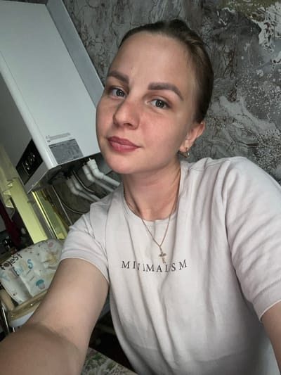 The image is a selfie taken indoors, likely in a residential setting in Balezino, Russia. The subject is a young woman with her hair pulled back and wearing a light-colored t-shirt with the word "MINIMALISM" printed on it in black letters, along with a small cross pendant necklace. She is looking at the camera with a neutral expression. Behind her, on the left side of the frame, is a white appliance, possibly a water heater or boiler, with visible pipes and wiring. The background on the right features a textured wall with a grey and white marbled pattern. A small part of a baby chair or stroller is visible on the far left, with a pattern of cartoon animals and the word "forest" visible.