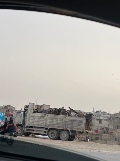 An overloaded truck is parked on a street lined with buildings, possibly apartments, against a hazy sky. The truck's bed is filled with scrap metal and other debris. People are visible near the truck's front. The image is shot from inside a vehicle, as a car frame is on the left side of the image.