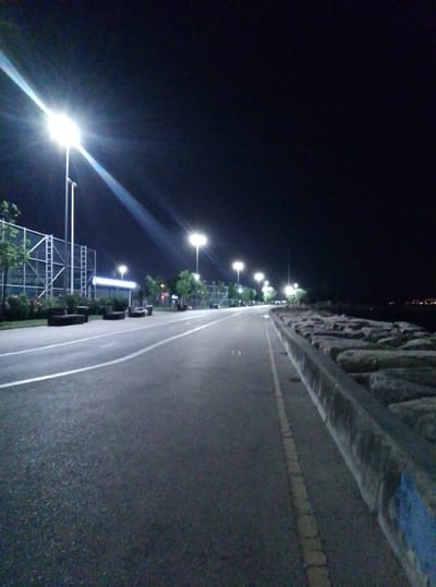 An empty paved walkway runs alongside a body of water on the right. The walkway has a white line marking its left edge and a yellow dashed line marking its right edge. A concrete barrier with large rocks is situated between the walkway and the water. On the left side of the walkway, there is a fence and several streetlights illuminate the area. The scene is dark, suggesting it is nighttime, with the sky appearing black. The location is the Moda Coastal Park in Kadıköy, Istanbul, Turkey, a popular spot for evening strolls.