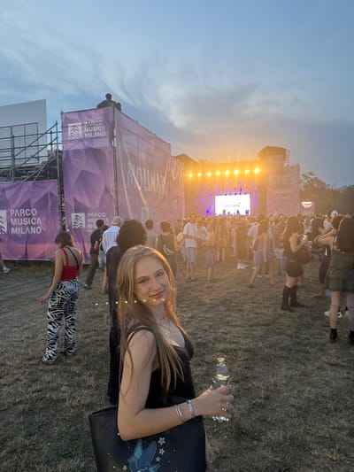 The image depicts an outdoor event, likely a concert or festival, at Parco Musica Milano in Segrate, Italy. The sky is overcast with patches of sunlight breaking through, casting a warm glow on the scene. In the foreground, a young woman with blonde hair is smiling at the camera, holding a clear plastic bottle of water in her right hand. She is wearing a black tank top and a silver bracelet. To her left, a person with dark hair is partially visible, wearing a black top and carrying a black bag adorned with a blue bird and stars.
In the background, a large stage with bright lights and a screen displaying text is set up. Several banners with the "Parco Musica Milano" logo are visible, as well as scaffolding structures. A crowd of people, dressed in casual attire, are gathered in front of the stage, some appearing to be listening to a performance. The ground is covered in dry grass. The overall atmosphere suggests a lively, social event taking place in the late afternoon or early evening.