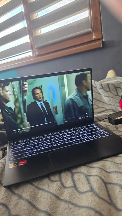 A black laptop is open on a bed with a gray patterned bedspread, with a window frame visible in the background. A film or show is playing on the laptop screen. The scene on the screen depicts two men, one in a blue shirt and another in a suit, with a blue light effect around them. The laptop keyboard has a white backlight. The window has blinds. The city of Valchi Dol, Bulgaria is not visible in the image.